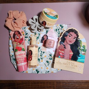 May include: A collection of Disney Moana themed beauty products including a mango scented bubble bath tube, a body scrub jar, a bottle of bath salts, a coconut scented lip mask, and a facial sheet mask, alongside a peach scrunchie and a gray hair claw.