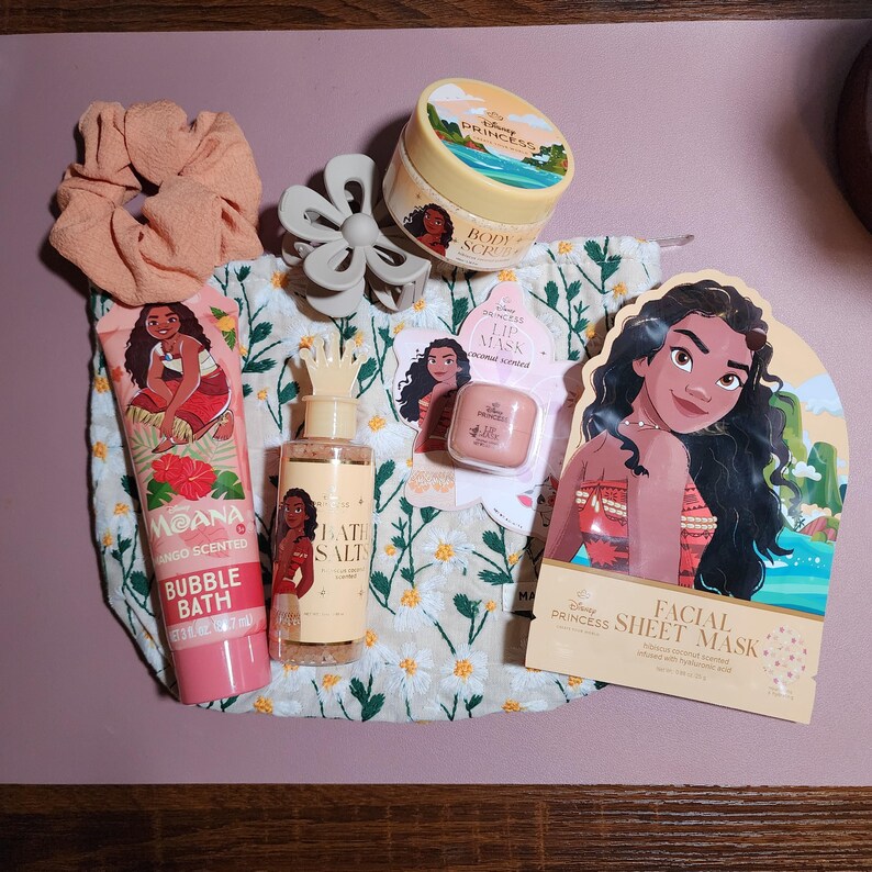 May include: A collection of Disney Moana themed beauty products including a mango scented bubble bath tube, a body scrub jar, a bottle of bath salts, a coconut scented lip mask, and a facial sheet mask, alongside a peach scrunchie and a gray hair claw.