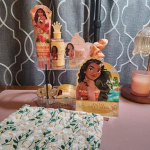 May include: A collection of Moana-themed bath and skincare products including a bubble bath tube, bath salts with a crown cap, a body scrub jar, and a facial sheet mask, all arranged on a clear two-tier stand next to a white floral embroidered cloth.