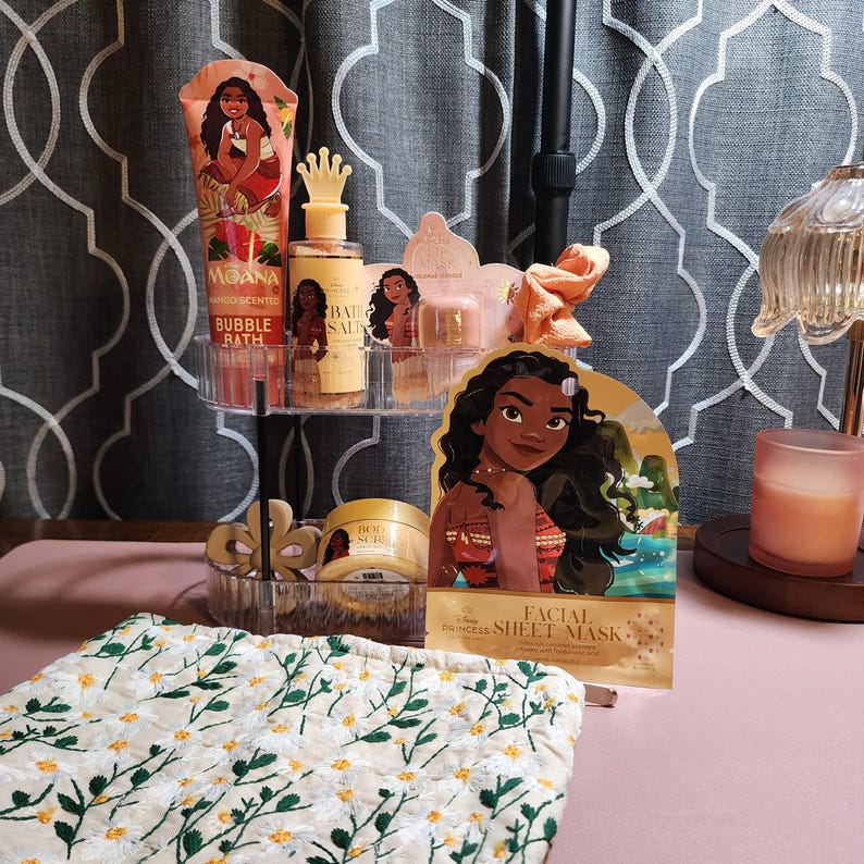 May include: A collection of Moana-themed bath and beauty products including a bubble bath tube, a bath salt bottle with a crown cap, a body scrub jar, and a facial sheet mask, all arranged on a two-tier clear stand with a floral patterned fabric.