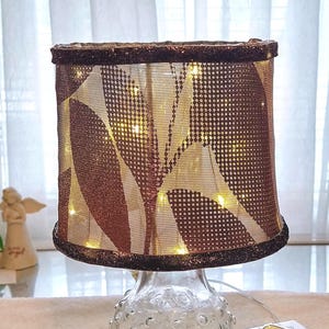 May include: A cylindrical lamp shade with a metallic copper and brown leaf pattern, featuring a dark glittery trim at the top and bottom edges. The shade is illuminated from within by small warm white LED string lights and sits on a clear glass base.