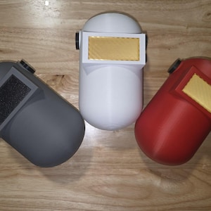 May include: Three welding helmets in gray, white, and red are displayed on a wooden surface. Each helmet has a rectangular visor with a gold-colored insert and a black adjustment knob. The helmets are designed for protection during welding.