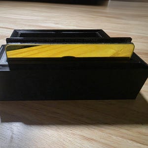 May include: A black rectangular box with a yellow and wood-grain top. The top section has a recessed area. The box is sitting on a wooden surface. The text on the top is blurry.