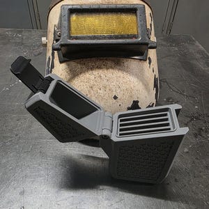 May include: An aged welding helmet with a worn, off-white finish and a rectangular gold-tinted lens. A gray, open, 3D-printed accessory with a honeycomb pattern and ventilation slats is in front of the helmet.