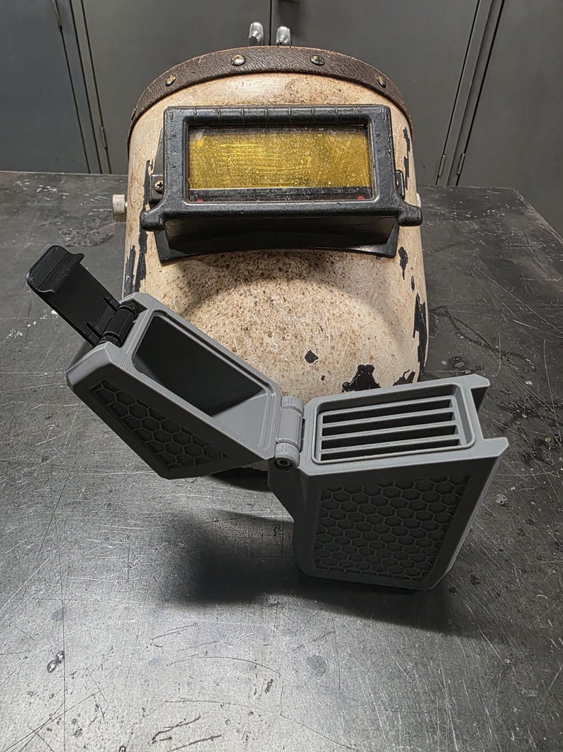 May include: An aged welding helmet with a worn, off-white finish and a rectangular gold-tinted lens. A gray, open, 3D-printed accessory with a honeycomb pattern and ventilation slats is in front of the helmet.