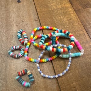 May include: A collection of colorful beaded bracelets and earrings. The bracelets are made with various colors of beads, including blue, green, pink, orange, and yellow. The earrings are made with a similar color scheme and feature a small pearl accent.