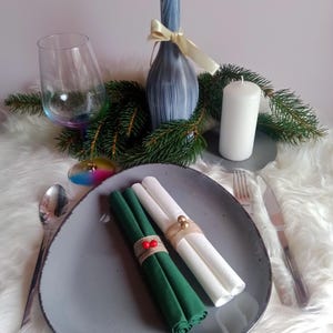 Può includere: Due tovaglioli arrotolati, uno verde e uno bianco, sono legati con spago e decorati con bacche rosse. I tovaglioli sono su un piatto grigio con una forchetta e un coltello accanto.