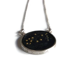 May include: A silver necklace with a round pendant featuring the Gemini constellation in gold beads on a black background. The pendant is engraved with the word "GEMINI".