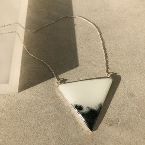 Puede incluir: Un collar de cadena de plata con un colgante triangular blanco con vetas negras. El colgante es de forma geom&eacute;trica con un acabado liso y brillante.