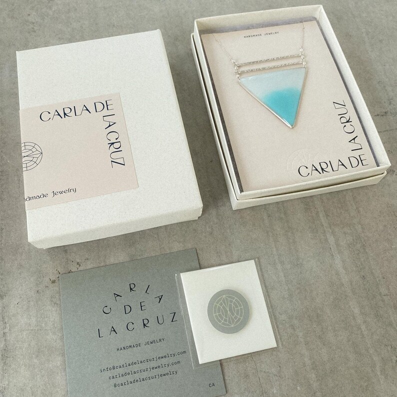 Puede incluir: Una caja blanca con el texto "CARLA DE LA CRUZ Handmade Jewelry" en la parte delantera. La caja contiene un collar de plata con un colgante triangular turquesa y blanco. El collar tambi&eacute;n se muestra en una caja blanca separada con el texto "CARLA DE LA CRUZ Handmade Jewelry" en la parte delantera.