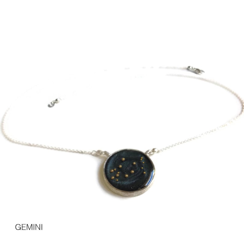 May include: A silver chain necklace with a round pendant featuring a dark blue background with gold glitter and the Gemini constellation.