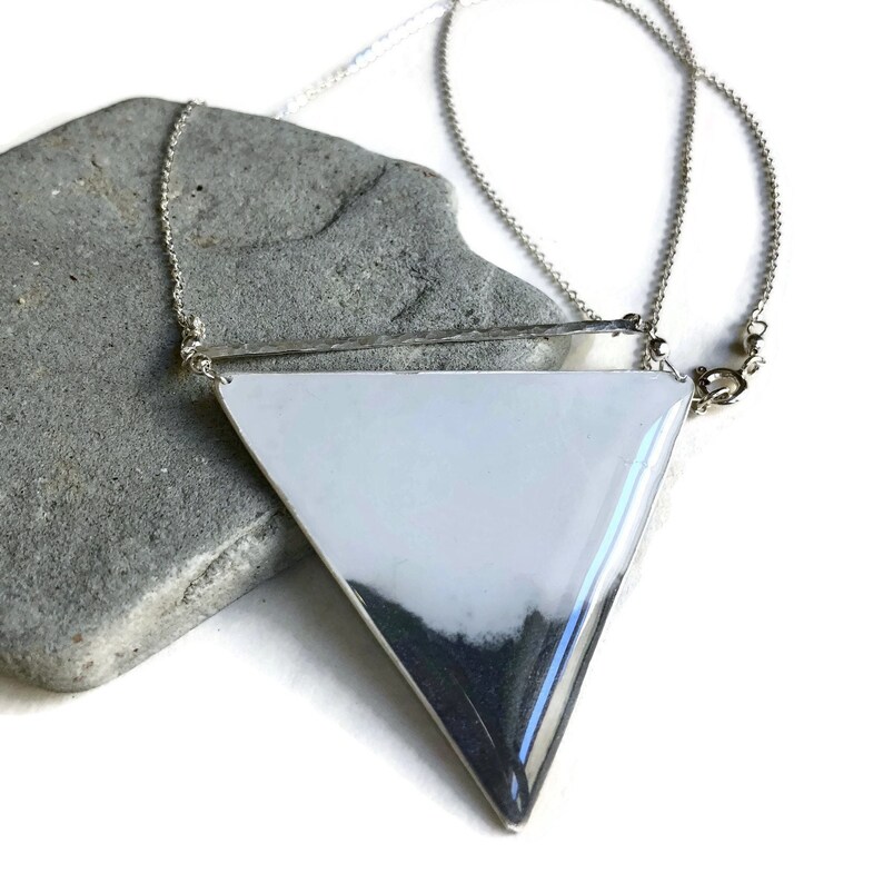 Puede incluir: Un collar con un colgante de tri&aacute;ngulo plateado y una cadena delicada. El colgante es grande y geom&eacute;trico, con un acabado liso y pulido.