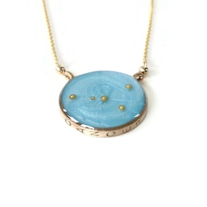 May include: A gold chain necklace with a round blue pendant featuring a constellation of gold stars. The pendant is inscribed with the letters "O M E G A Z".