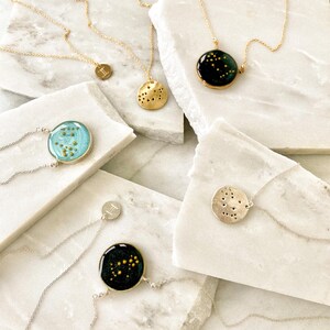 May include: Five gold necklaces with different zodiac constellation pendants. Each pendant features a different constellation design with gold glitter embedded in clear resin. The pendants are round and have a silver backing.