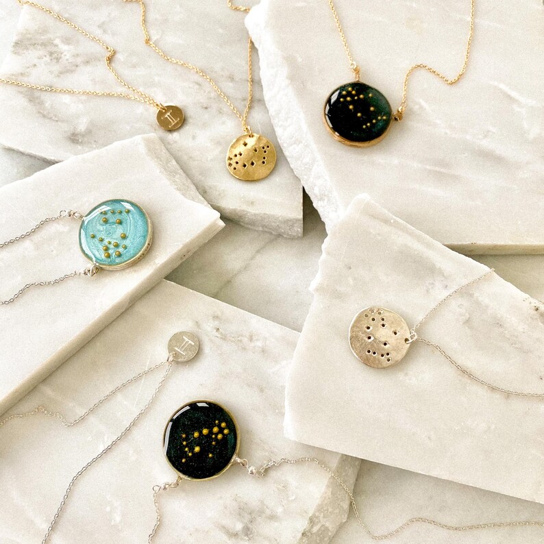 May include: Five gold necklaces with different zodiac constellation pendants. Each pendant features a different constellation design with gold glitter embedded in clear resin. The pendants are round and have a silver backing.