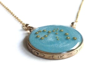 May include: A gold chain necklace with a round pendant featuring a blue background and gold dots arranged in a constellation pattern. The pendant has the word "GOD" inscribed around the edge.