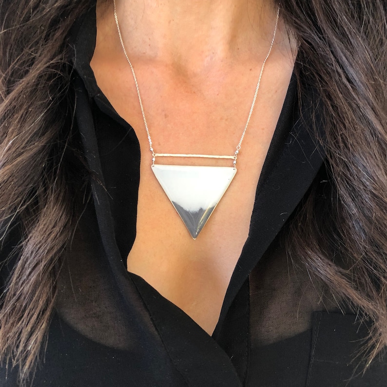 Puede incluir: Un collar con un colgante de tri&aacute;ngulo plateado con una barra fina en la parte superior. El colgante es grande y cuelga de una cadena delicada.