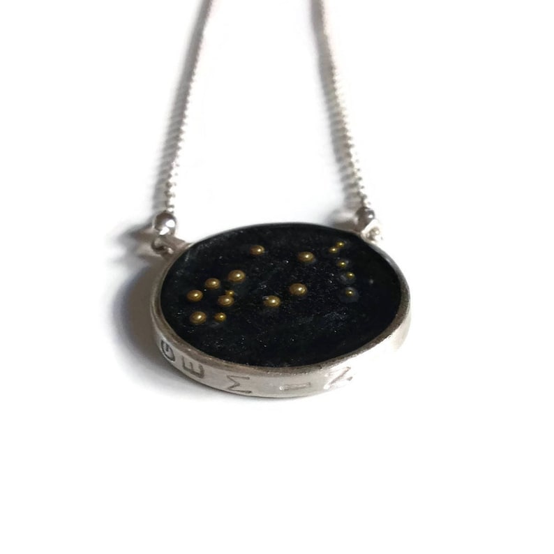 May include: A silver necklace with a round pendant featuring the Gemini constellation in gold beads on a black background. The pendant is engraved with the word "GEMINI".