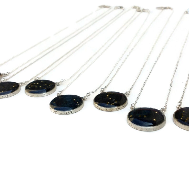 May include: A set of silver necklaces with round pendants featuring constellations and zodiac signs. The pendants are black with gold glitter and have the name of the constellation inscribed around the edge.