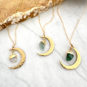 May include: Three gold necklaces with crescent moon pendants and different colored crystal charms. The charms are a clear quartz, a green aquamarine, and a green emerald.