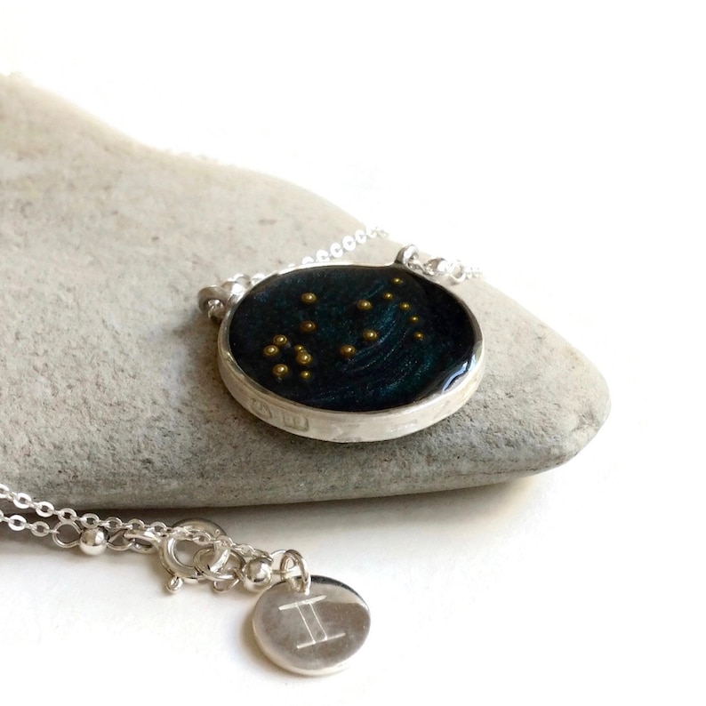 May include: A silver necklace with a round pendant featuring a dark blue background with gold stars representing the Gemini constellation. A smaller silver charm with the Gemini symbol hangs below the pendant.