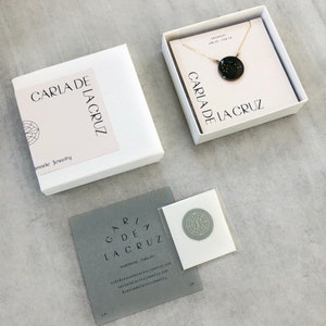 May include: A white box with the text "CARLA DE LA CRUZ" and "Handmade Jewelry" on it. A smaller white box with a gold chain and a black pendant with gold stars inside. The pendant has the text "CARLA DE LA CRUZ" on it. A gray card with the text "CARLA DE LA CRUZ" and contact information.