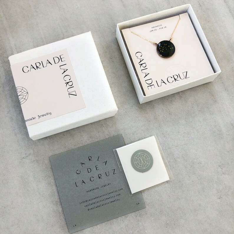 May include: A white box with the text "CARLA DE LA CRUZ" and "Handmade Jewelry" on it. A smaller white box with a gold chain and a black pendant with gold stars inside. The pendant has the text "CARLA DE LA CRUZ" on it. A gray card with the text "CARLA DE LA CRUZ" and contact information.