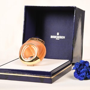 Op de afbeelding: Een gouden en oranje parfumfles met een ringontwerp, in een blauwe doos met de tekst "BOUCHERON Paris" op de deksel. Een blauwe bloem is op de voorgrond.