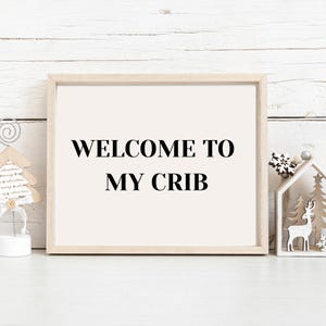 Può includere: Una stampa incorniciata con la scritta "WELCOME TO MY CRIB" in nero. La cornice è in legno chiaro. Ornamenti natalizi decorativi sono a destra e a sinistra della cornice.