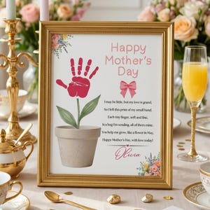 Puede incluir: Un marco dorado con una impresión para el Día de la Madre. La obra de arte presenta una flor con una huella de mano roja, el texto "Happy Mother's Day" y un poema. También se incluyen detalles florales y el nombre "Olivia".