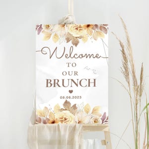 May include: A white sign on an easel with the words "Welcome to our Brunch" in brown script. The sign is decorated with floral elements in shades of yellow, brown, and white. The date "08.06.2023" is printed below the word "Brunch".