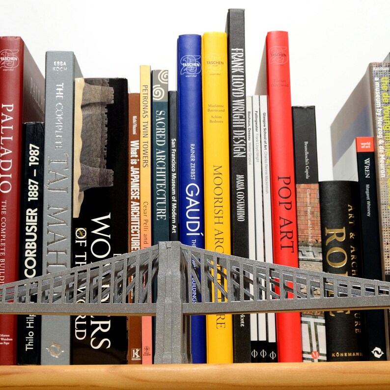 Lifestyle image features a partial view of an assembled Brooklyn Bridge model. Model of this suspension bridge is made out of silvery cardstock parts, placed on a wooden shelf in front of art books. The view features one pylon, the deck and cables.