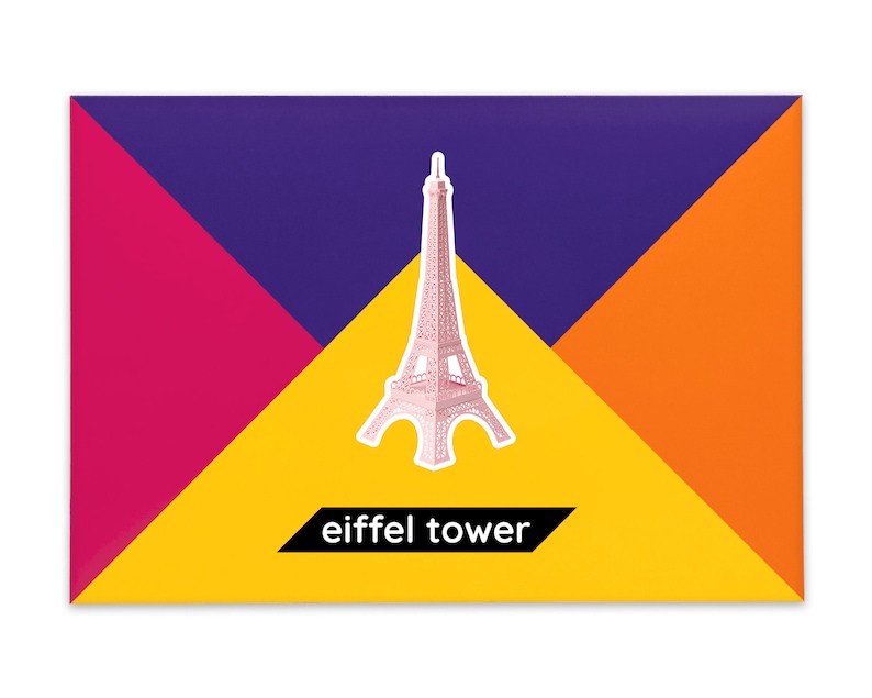 Product packaging for the Eiffel Tower paper model kit. A recangular bright coloured cardstock envelope features four large triangles in yellow, magenta, purple and orange with an image of a finished model at the centre and the product title below.
