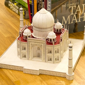 Lifestyle image shows the assembled 3d paper model of Taj Mahal on a solid wood surface with books behind it. The model features the central dome, four kiosk domes, four minarets, vaulted structures and panels.