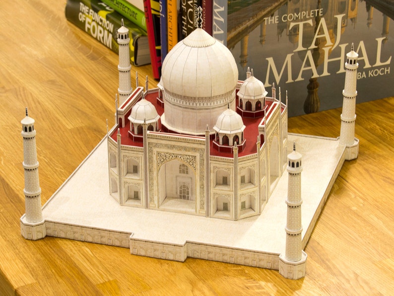 Lifestyle image shows the assembled 3d paper model of Taj Mahal on a solid wood surface with books behind it. The model features the central dome, four kiosk domes, four minarets, vaulted structures and panels.