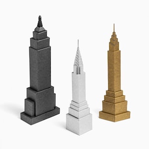 May include: Three miniature skyscraper models in shades of grey, silver, and gold. The buildings have a stepped design with detailed architectural features. The models are set against a white background.