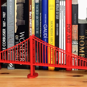 Lifestyle image features a partial view of an assembled Golden Gate bridge model. Model of this suspension bridge is made out of red cardstock parts, placed on a wooden shelf in front of art books. The view features one pylon, the deck and cables.