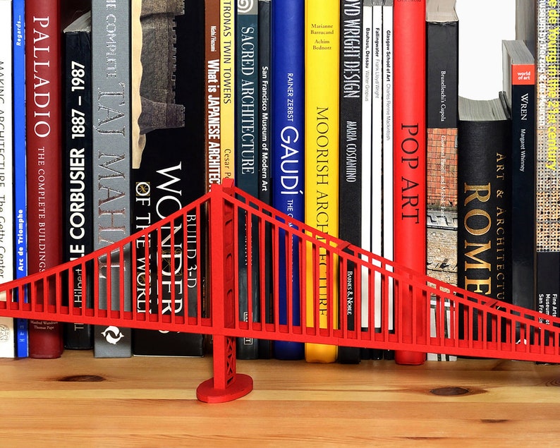 Lifestyle image features a partial view of an assembled Golden Gate bridge model. Model of this suspension bridge is made out of red cardstock parts, placed on a wooden shelf in front of art books. The view features one pylon, the deck and cables.