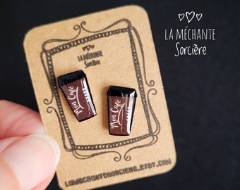 May include: A pair of coffee cup-shaped earrings with the words "Mon Cafe" printed on them. The earrings are brown with black tops and are displayed on a brown card with decorative borders and the text "LA MÉCHANTE Sorcière".