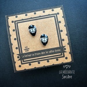 May include: A pair of black and white hockey mask earrings. The earrings are on a brown card with the text "Le bonheur se trouve dans les petites choses" and "LA MÉCHANTE Sorcière".