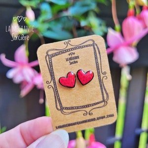 May include: A pair of red heart-shaped earrings with black outlines on a brown card with the text "La M&eacute;chante Sorci&egrave;re" and a decorative border.