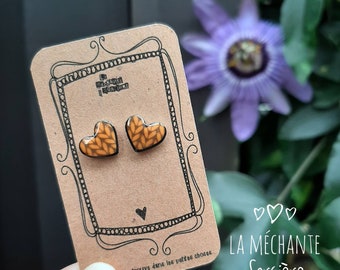 May include: A pair of heart-shaped earrings with a knitted pattern in shades of orange and brown. The earrings are displayed on a brown card with the text "La Méchante Sorcière" and "Le trouve dans les petites choses".