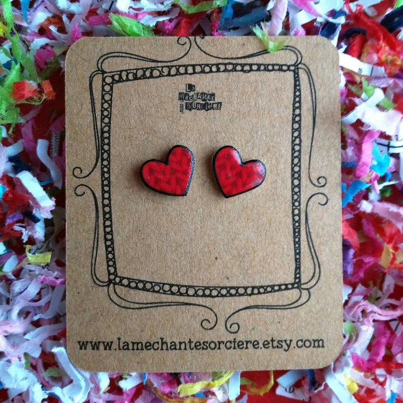 May include: A pair of red heart-shaped earrings with a black and white patterned design. The earrings are on a brown card with the text "www.lamechantesorciere.etsy.com".