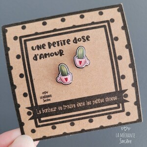 Puede incluir: Un par de pendientes con un peque&ntilde;o cactus en una taza de t&eacute; rosa con un coraz&oacute;n en el lateral. Los pendientes est&aacute;n en una tarjeta con el texto "UNE PETITE dose D'AMOUR" y "Le bonheur se trouve dans les petites choses" en franc&eacute;s. La tarjeta tambi&eacute;n tiene el texto "LA M&Eacute;CHANTE Sorci&egrave;re" en franc&eacute;s.