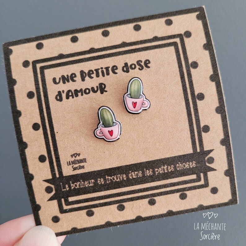 Puede incluir: Un par de pendientes con un peque&ntilde;o cactus en una taza de t&eacute; rosa con un coraz&oacute;n en el lateral. Los pendientes est&aacute;n en una tarjeta con el texto "UNE PETITE dose D'AMOUR" y "Le bonheur se trouve dans les petites choses" en franc&eacute;s. La tarjeta tambi&eacute;n tiene el texto "LA M&Eacute;CHANTE Sorci&egrave;re" en franc&eacute;s.