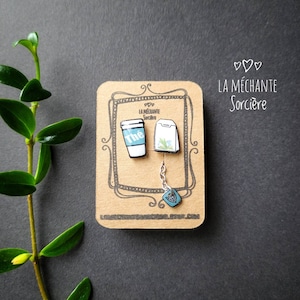 May include: A pair of earrings featuring a blue coffee cup with the word "the" on it and a blue tea bag with a white string. The earrings are displayed on a brown card with the text "La Méchante Sorcière" in white.