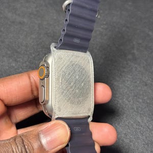 Puede incluir: Un reloj inteligente plateado con una correa azul oscuro. El reloj tiene una esfera naranja y una parte trasera texturizada. La correa está marcada con "49mm" y "S/M".