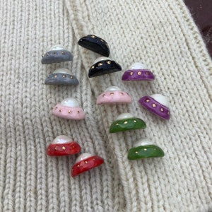 May include: A collection of 10 ceramic buttons in various colors, including pink, green, red, purple, and gray. Each button features a white pearl-like center and gold accents around the edge.
