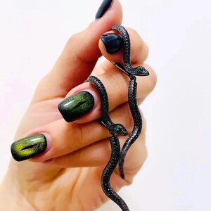 May include: A pair of black snake earrings with green gem eyes. The earrings are detailed with small, dark embellishments and are held up against a white background. The snake design is a unique and eye-catching jewelry piece.