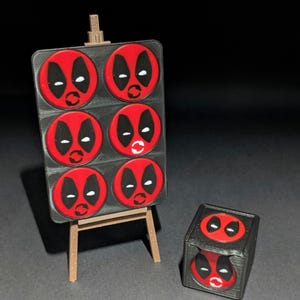Può includere: Un cavalletto in miniatura mostra sei disegni di volti di Deadpool rossi e neri. Un cubo abbinato con due volti di Deadpool è accanto al cavalletto. Gli articoli sono stampati in 3D e sono probabilmente oggetti da collezione o decorativi.